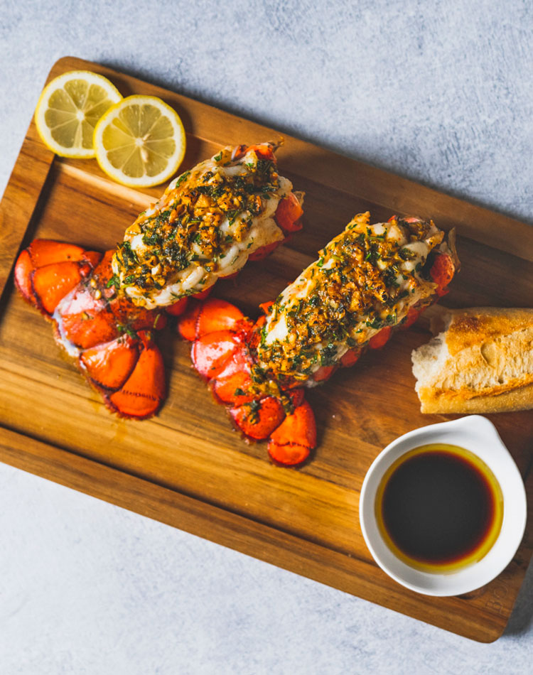 Wooden cutting board with lobster tails, lemon slices, and a small bowl of sauce on a light gray background.