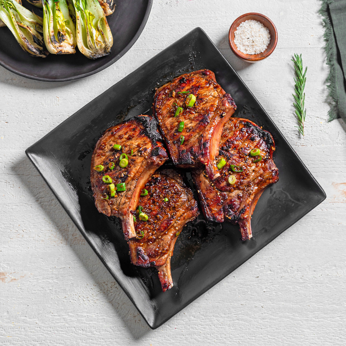 Grilled pork chops on a black plate with a side of grilled vegetables on a light gray background.