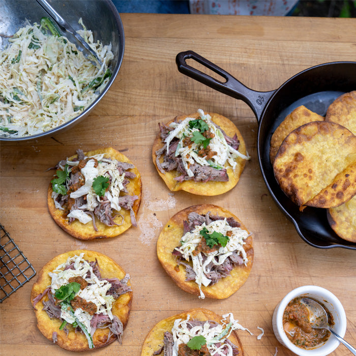 Wooden cutting board with pork tostadas, a skillet, and a bowl of ingredients.
