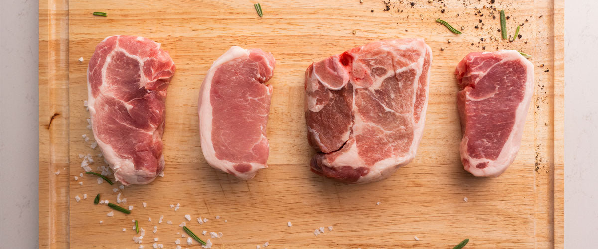 Four raw pork chops on a wooden cutting board with herbs and spices.