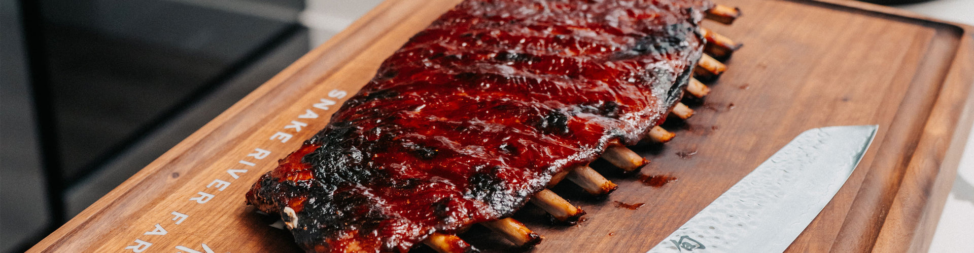 Grilled ribs on a wooden cutting board with a knife, on a kitchen counter.