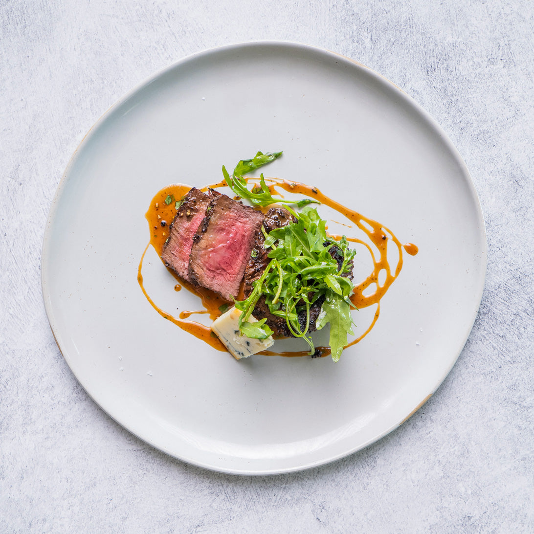 Plated dish with steak, greens, and sauce on a light gray background