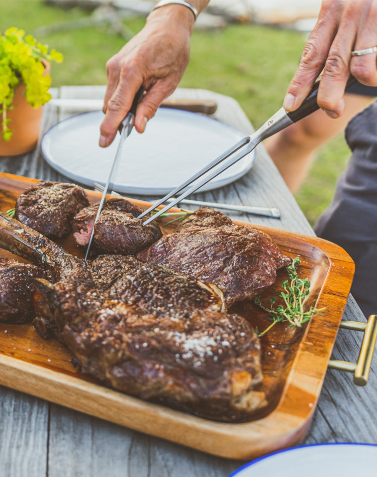 Grilled steaks on a wooden cutting board with tongs, outdoors.