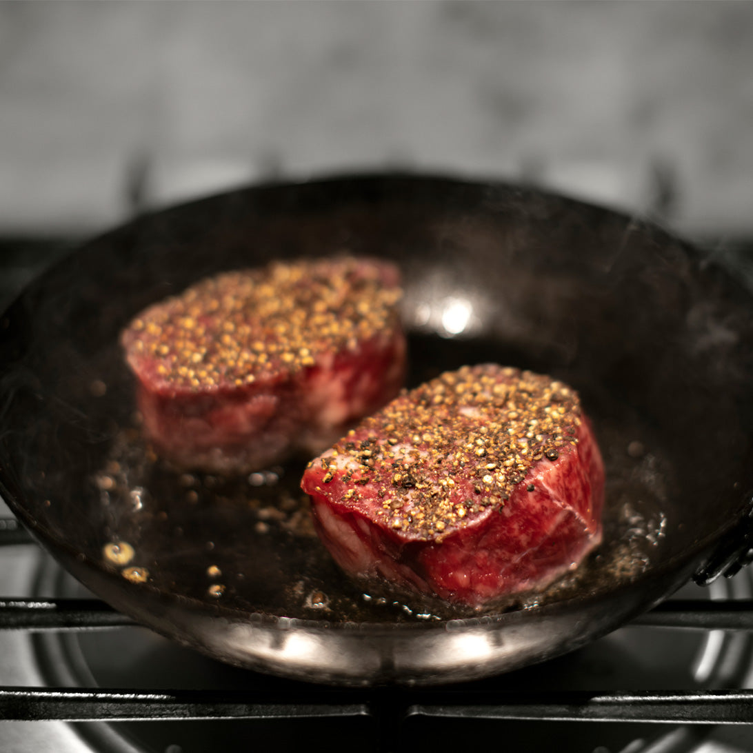 Two raw steaks seasoned with herbs and spices cooking in a black cast iron skillet.